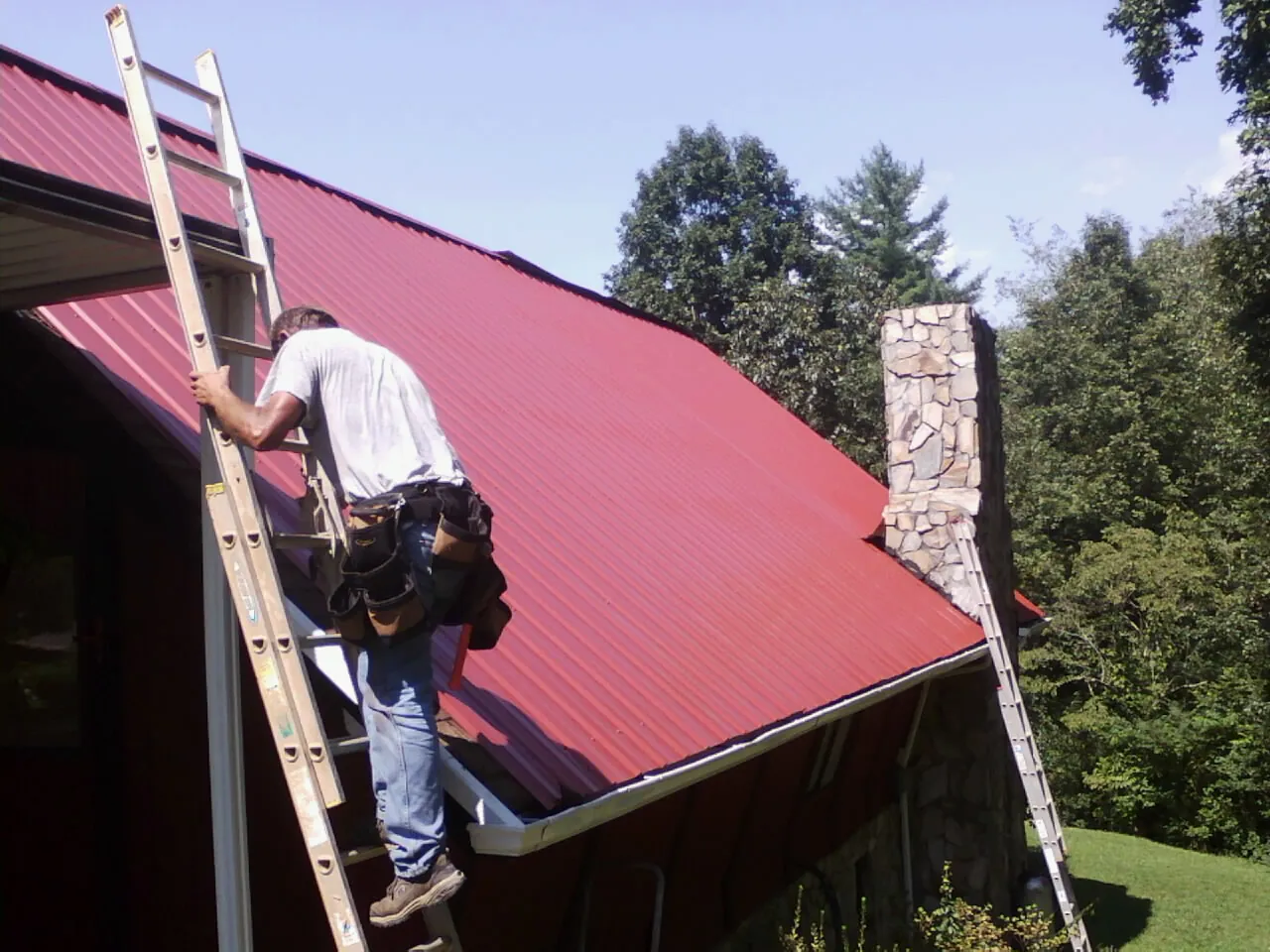Roofer on a ladder beside a red metal roof and stone chimney — Soffit & Fascia Repair by Roofing Brothers in Smiths Grove
